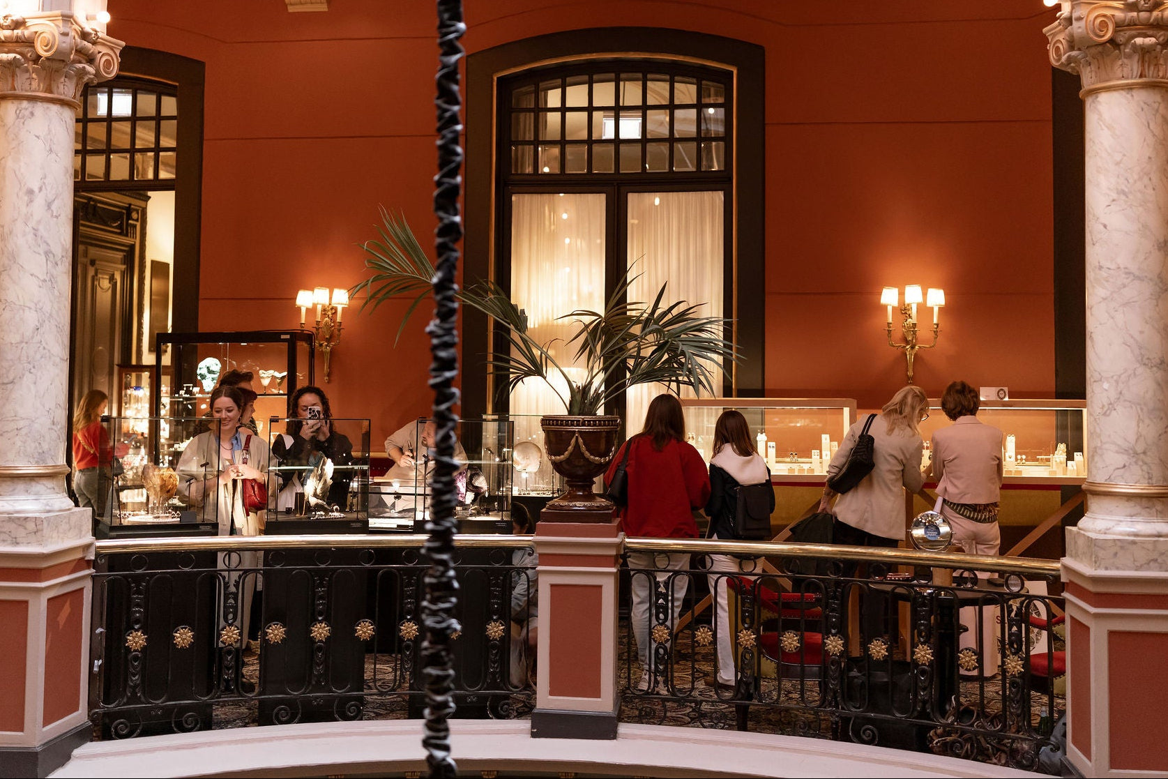 A wide interior scene of a grand hotel or casino lobby with tall marble columns, warm orange walls, arched windows, and several people standing and sitting near a bar or lounge area in the background.