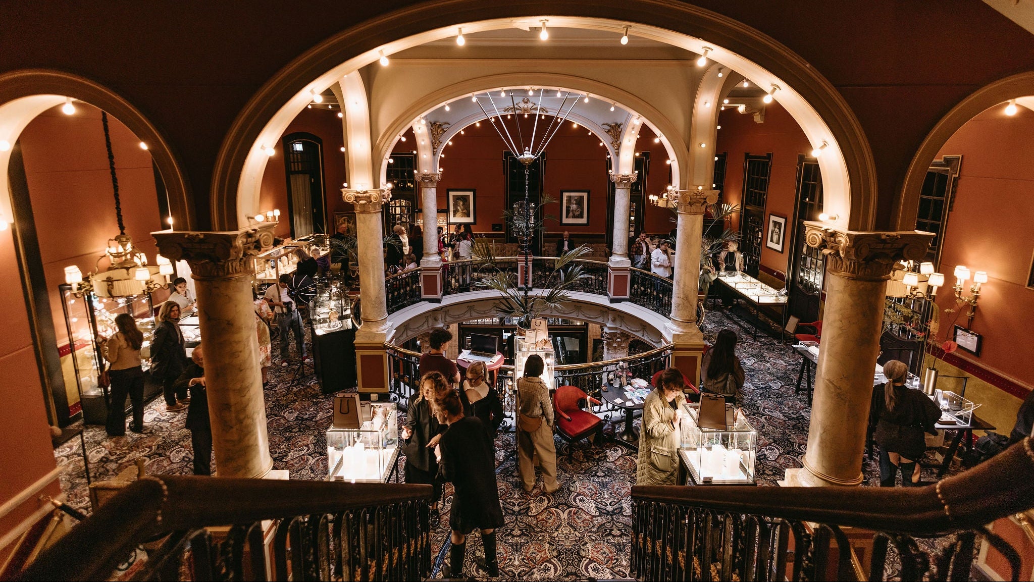 An elevated view of a grand, historic interior hall with arched ceilings, warm red-brown walls, ornate columns, chandeliers, and many people gathered around display tables on the lower level.
